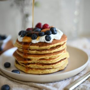 Stack of pancakes with blueberries and raspberries topped with syrup and whipped cream on a plate.